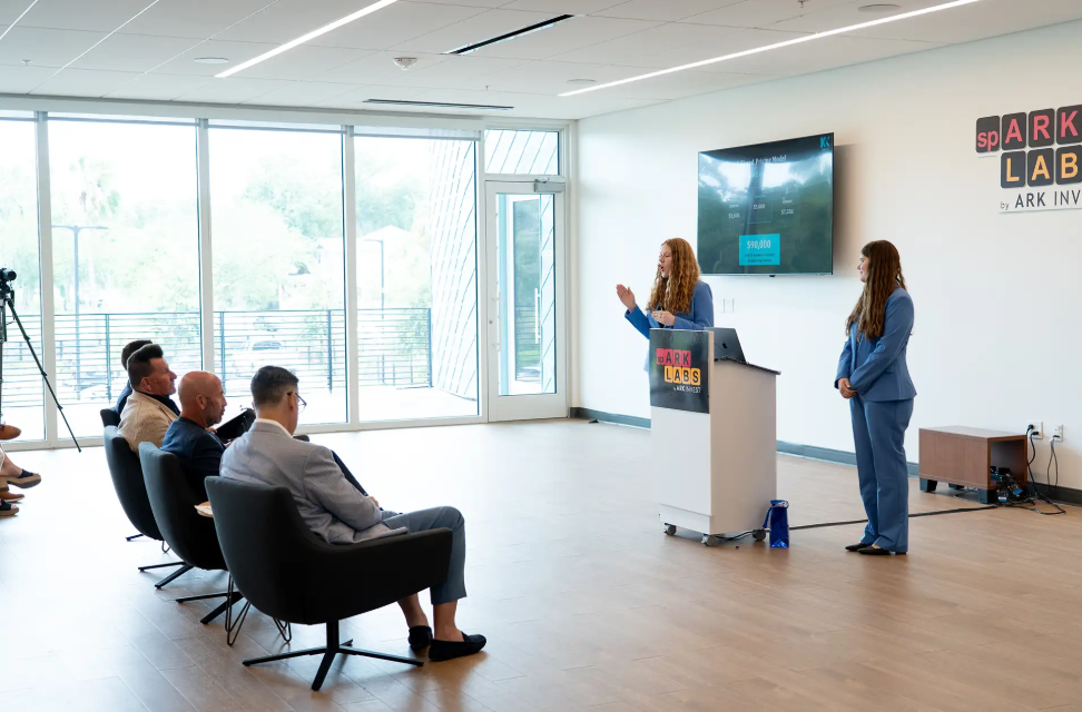 Presenting at a Spark Labs podium, with Kevin Harrington seated in the audience.