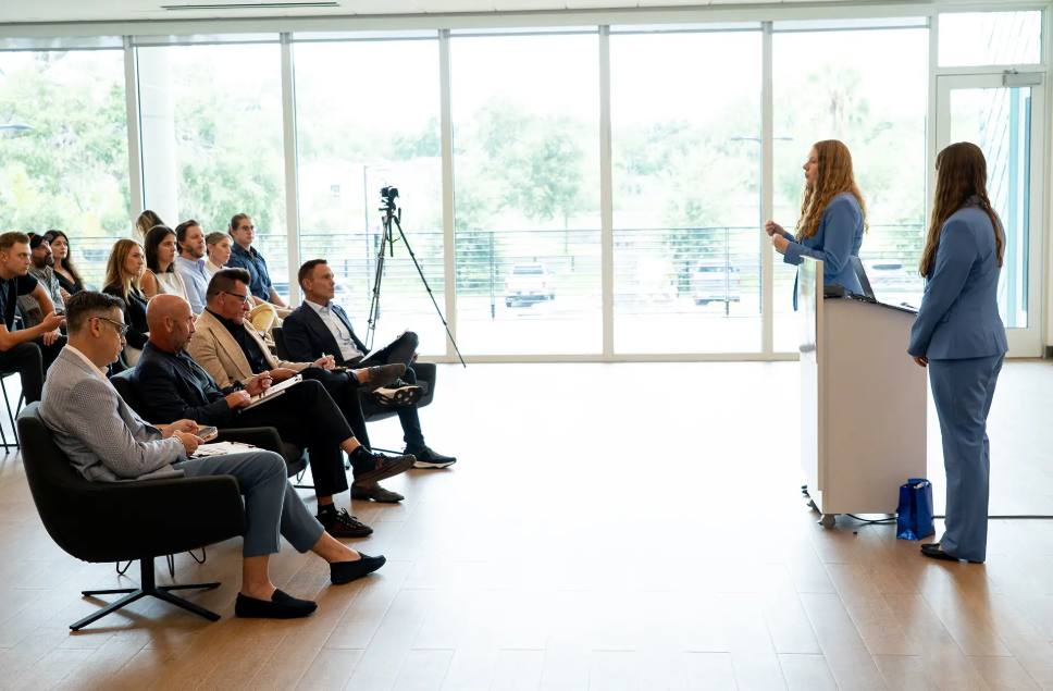 Presenting from a podium to a seated audience in a room with floor-to-ceiling windows.